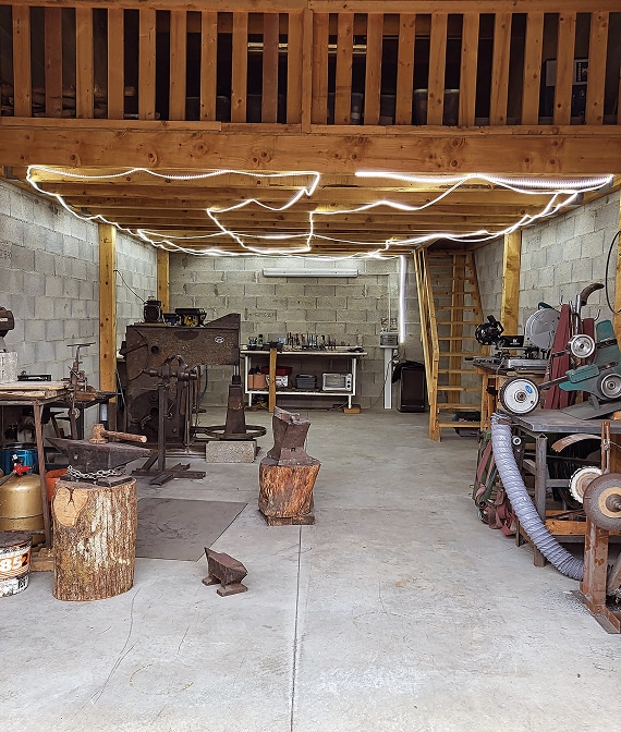 Forge industrielle : aménagement d'atelier avec mezzanine et LED. Atelier de forge industriel. Mezzanine en bois, murs en blocs de béton, sol en ciment. Enclumes sur billots et outils de travail du métal sous un éclairage LED.