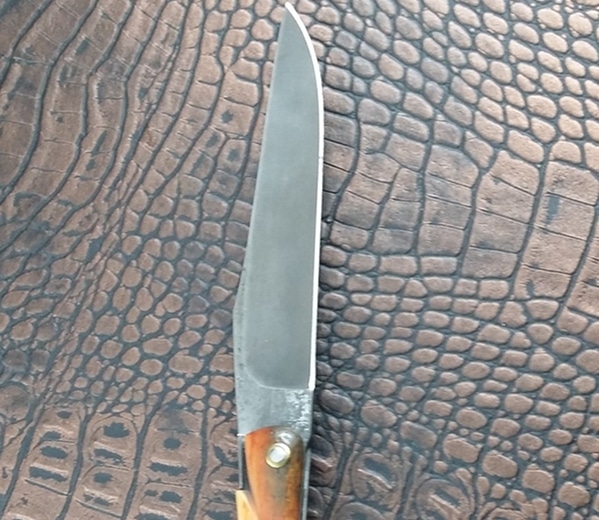 Lame de couteau pliant artisanal à dos droit, finition forge brute, contrastant avec le manche en bois et le fond texturé en cuir crocodile.