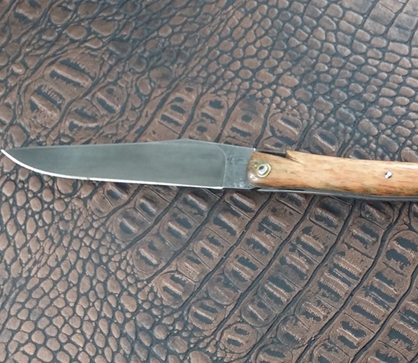 Couteau pliant à lame acier brut et manche en bois clair, posé sur un fond texturé brun de type cuir de crocodile.