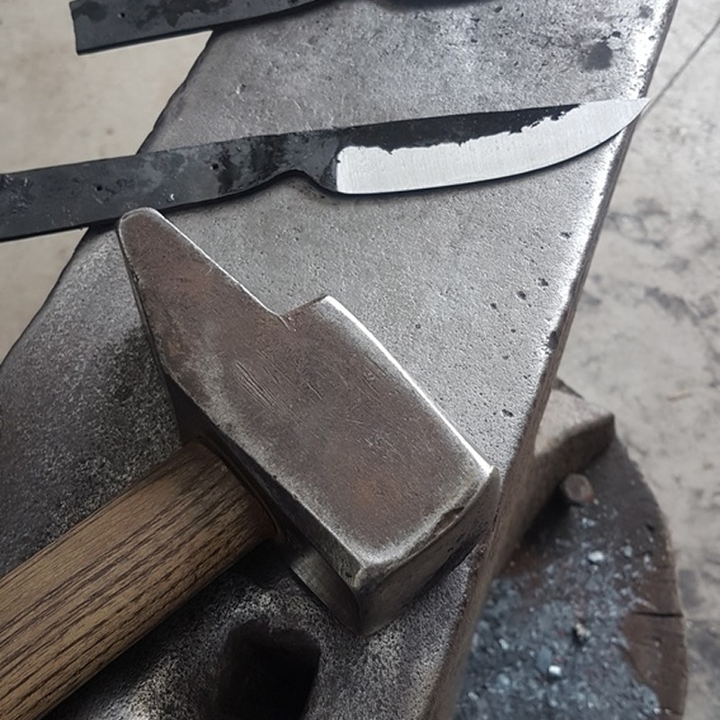 Marteau de forge et lame de couteau brute à tranchant poli reposant sur une enclume en acier texturé dans un atelier.